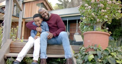 African american father hugging laughing son on porch steps of modern family home