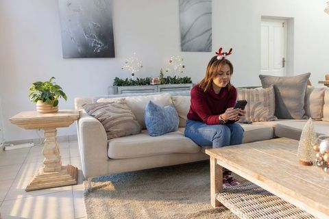 Asian woman sitting on sofa wearing reindeer antlers holding phone in cozy holiday room