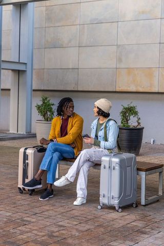 Diverse friends sitting with suitcases on urban bench, chatting while waiting at plaza