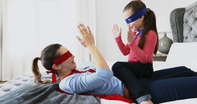 Happy caucasian mother and daughter in superhero masks and capes playing on bed. Fun, playing, dressing up, childhood, motherhood, togetherness, free time and domestic life, unaltered.