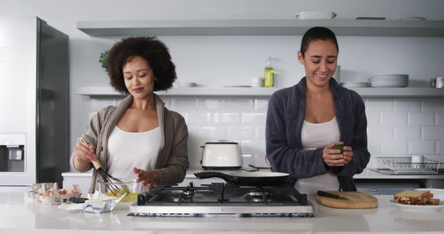 Two women are smiling and enjoying cooking together in a modern kitchen. One woman is beating eggs, while the other holds a green vegetable, both engaged in food preparation. Distinctive features include a gas stove, white subway tiles, and sleek countertops. This can be used for articles, advertisements, or social media posts focusing on home living, friendship, diverse communities, and culinary activities.