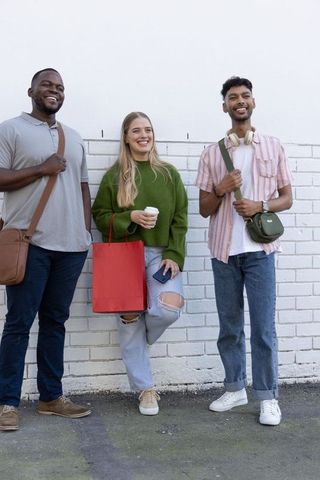 Diverse friends leaning against white brick wall smiling, holding coffee and shopping bag