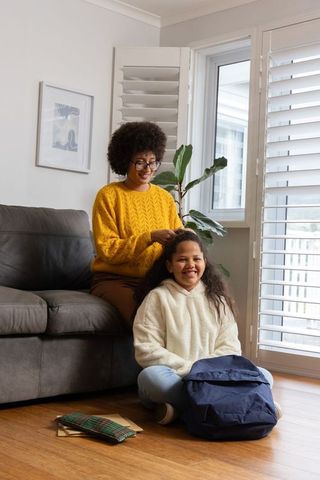Mother styling daughter hair in living room during cheerful back-to-school morning routine