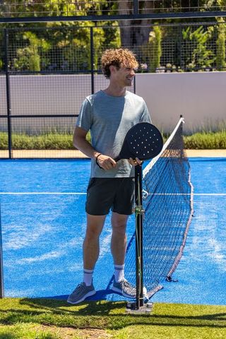 Young man holding padel racket smiling on blue outdoor court during sunny leisure match