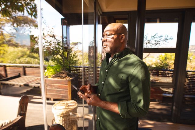 Senior African American man standing by window, holding smartphone, looking thoughtful. Ideal for use in articles about retirement, senior lifestyle, technology use among older adults, and moments of contemplation. Suitable for promoting products or services related to senior living, mobile technology, and home design.