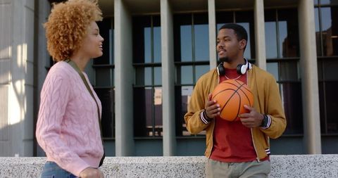 Young friends talking on campus plaza holding basketball wearing headphones outdoor casual