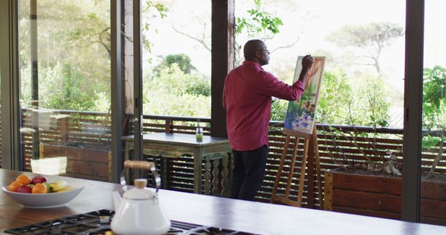 Happy african american senior man standing on sunny balcony painting picture of the view. retirement lifestyle, spending time alone at home.