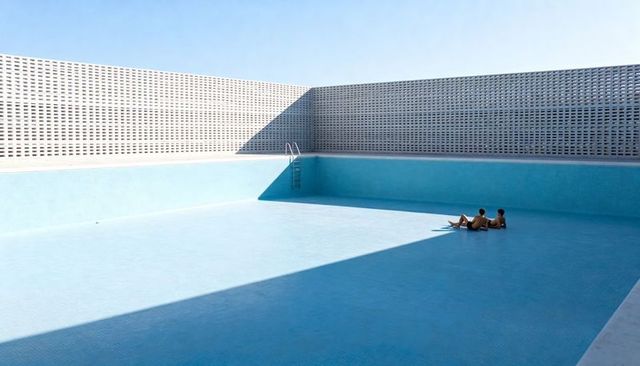 Minimal rooftop pool with two people reclining on empty blue-tiled floor under geometric shadow