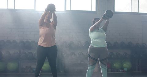 Two diverse women lifting kettlebells overhead in backlit gym for strength training
