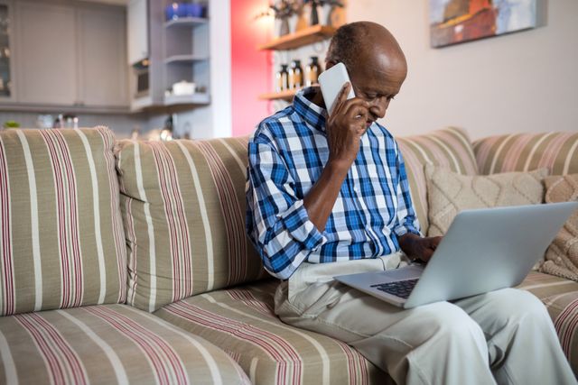 Senior man sitting on a striped couch in a cozy living room, talking on the phone while using a laptop. Ideal for illustrating concepts related to remote work, technology use among seniors, home office setups, and modern communication.