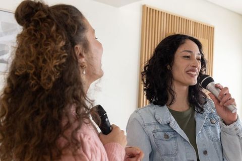 Female friends singing duet in casual home studio holding microphones near acoustic panel