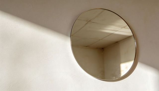 Round brass mirror reflecting tiled ceiling, warm diagonal sunlight casting soft shadows on textured