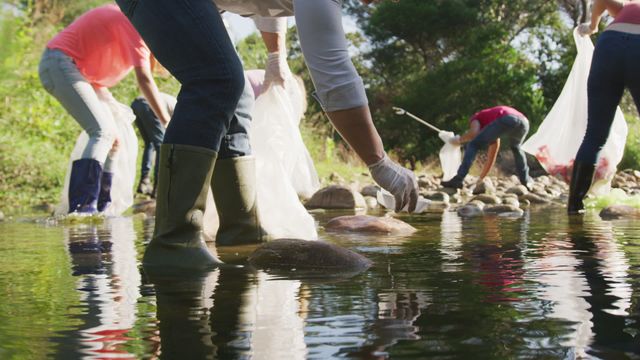 Conservation volunteers gather beside a rural river under the sun, cleaning up waste while equipped with gloves and boots. Ideal for use in materials promoting environmental awareness, community service, and ecological responsibility initiatives. Perfect for illustrating teamwork, unity, and outdoor activities focused on protecting nature.