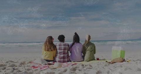 Four friends sitting on sandy beach watching ocean horizon with cooler and towel