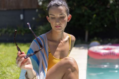 Contemplative woman sitting poolside in yellow swimsuit holding sunglasses near pink float