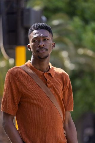 African american man standing on sunlit city sidewalk wearing rust polo and crossbody bag