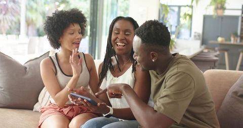 African American Friends Laughing and Sharing Smartphone on Living Room Sofa