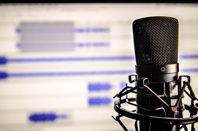 Close-up view of a professional microphone with a blurred audio editing software interface in the background. Ideal for use in articles or visuals related to podcasting, audio recording, voiceover work, broadcasting, or music production. This stock image is perfect for media production websites, blogs about sound engineering, and banners for recording studio advertisements.