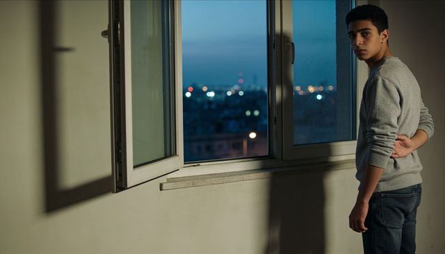 Teen standing at open window looking out over city at dusk with dramatic shadow