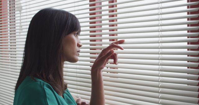 A woman wearing a green shirt looking through window blinds with a thoughtful expression. Ideal for illustrating themes of contemplation, solitude, and introspection in personal blog posts, mental health awareness articles, or lifestyle content.