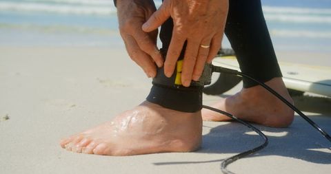 Senior surfer securing leash beside surfboard on sandy beach