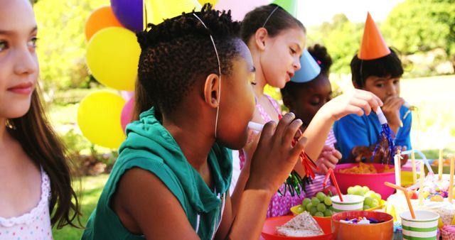 A diverse group of children enjoy a festive outdoor birthday party, with colorful balloons and party hats adding to the cheerful atmosphere. They are gathered around a table filled with snacks, engaging in the joyous celebration of a special occasion.