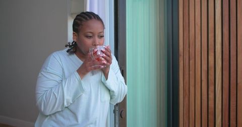 African american woman sipping tea at sliding glass door wearing loungewear morning calm and cozy
