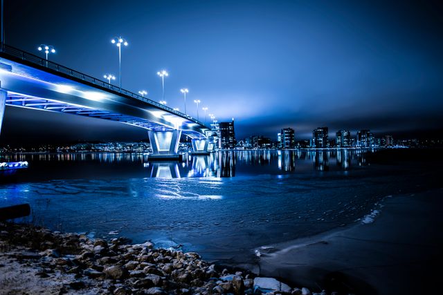 Spectacular view of an illuminated bridge extending over calm waters, reflecting bright city lights and creating a breathtaking urban landscape at night. Ideal for themes related to modern architecture, urban development, night scenes, travel destinations, and city skylines.