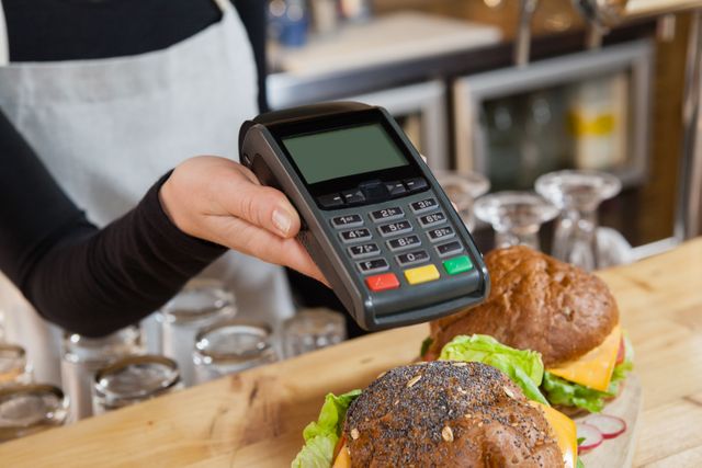 Cafe owner holding a credit card reader, ready to process a payment. Two burgers with fresh ingredients are on the counter. Ideal for illustrating small business operations, contactless payment methods, and customer service in a food establishment.