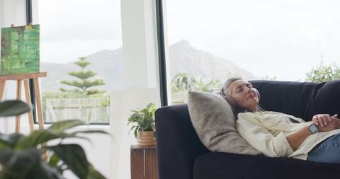 Mature woman relaxing on sofa with headphones and smartwatch in art-filled living room