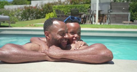 African american father and teen son leaning on pool edge smiling in backyard summer