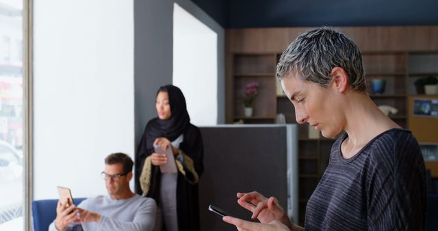 Diverse group of professionals engaging with smartphones in a modern office. One woman wearing a hijab stands in the background, while a man in glasses and another woman with short, grey hair focus on their devices. Ideal for use in projects related to workplace communication, technology integration, business environments, and modern office settings.