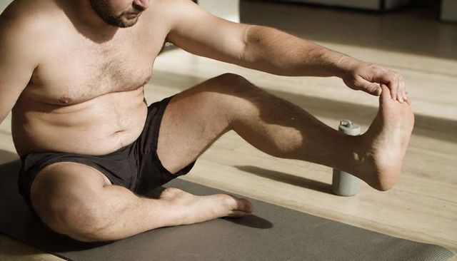 Shirtless man performing seated hamstring stretch on mat at home with water bottle