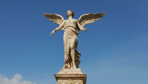 Neoclassical angel statue standing on ornate pedestal against clear blue sky
