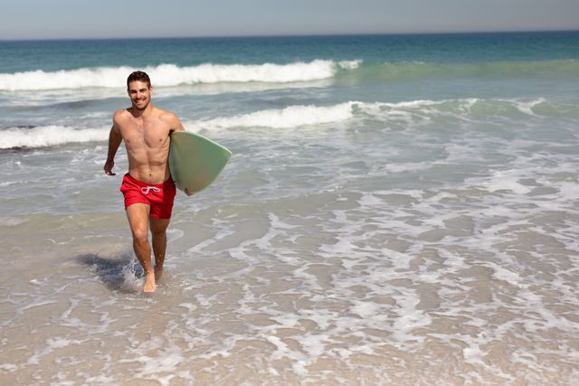 Front view of shirtless man with surfboard walking on beach in the sunshine