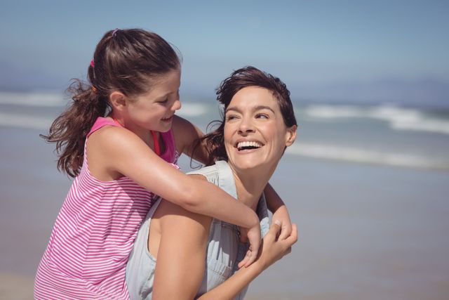 Happy mother piggybacking daughter at beach during sunny day
