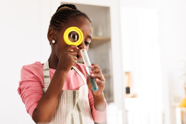 Young African American girl engaging in a science experiment at home, using a magnifying glass to examine a test tube. Ideal for educational content, STEM promotions, children's learning materials, and science-related advertisements.