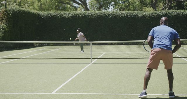 Two men playing tennis on an outdoor court during a sunny day. One man is serving the ball while the other prepares to return it. Ideal for promoting sports activities, outdoor events, active lifestyle benefits, and sports apparel.