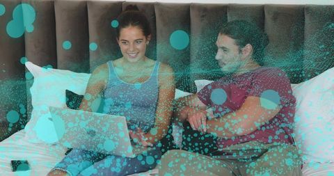 Couple relaxing on bed sharing laptop with teal abstract dot overlay