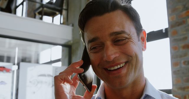 Businessman smiling while talking on phone in bright modern office. Ideal for projects related to business communication, professional services, and workplace productivity. Suitable for use in websites, advertisements, and corporate materials.