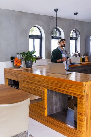 Indian man holding mug by laptop at wood kitchen island in cozy rustic contemporary space