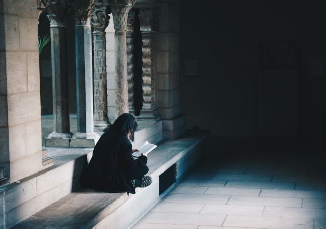 This scene captures a person engaged in reading within historic architecture, sitting alone on a stone bench. The image evokes feelings of solitude and tranquility, ideal for use in educational and historical content, promoting knowledge, contemplation, and study in a peaceful environment. This can be used in articles or websites focusing on historical research, libraries, and places of learning.