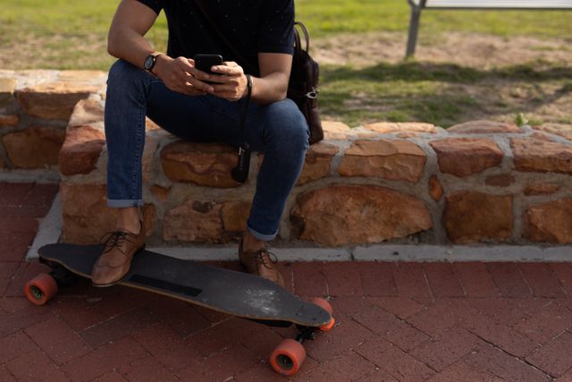 Front view mid section of a biracial man enjoying free time in nature on a sunny day, sitting on the wall with one feet on long board, using a smartphone. 