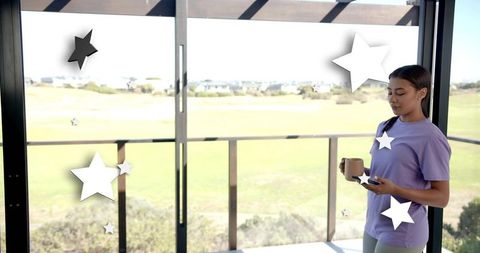 Young woman holding mug checking phone on sunny balcony with star stickers