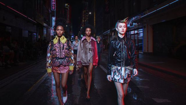 Models walking neon-lit wet city street wearing bomber jackets, skirts, fishnet stockings