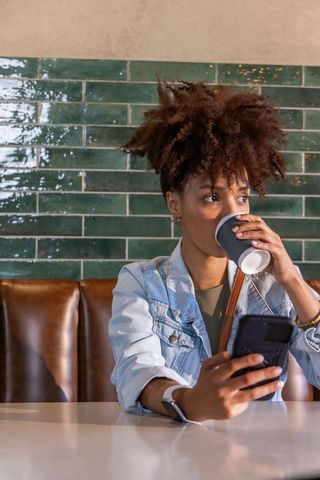 African american woman sipping coffee and checking phone in cafe booth with denim jacket