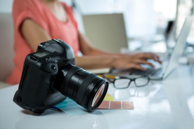 Digital camera placed on a table in a creative studio with a person working on a laptop in the background. Ideal for use in articles or advertisements related to photography, creative workspaces, professional equipment, or technology in the workplace.