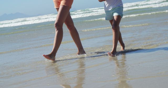 Capturing a moment of family bonding, this image shows a mother and her child walking barefoot along a sunny beach. Perfect for promoting family travel, beach vacations, and outdoor activities. Useful for advertisements targeting family-oriented products or travel destination promotions.