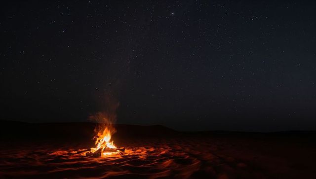 Campfire flickering on desert dunes under star-filled night sky with warm glowing embers