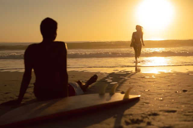 Woman running with surfboard while man relaxing on the beach during sunset